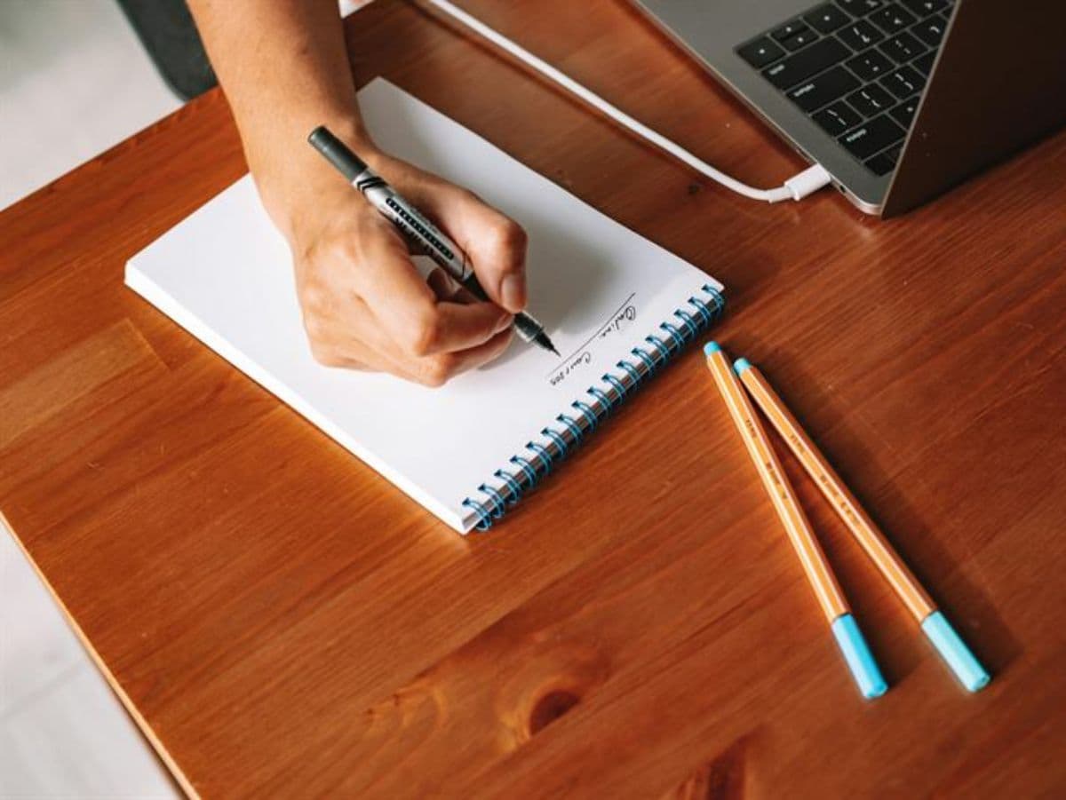 Mano escribiendo en un cuaderno junto a un portátil, preparando la documentación de un trámite en España